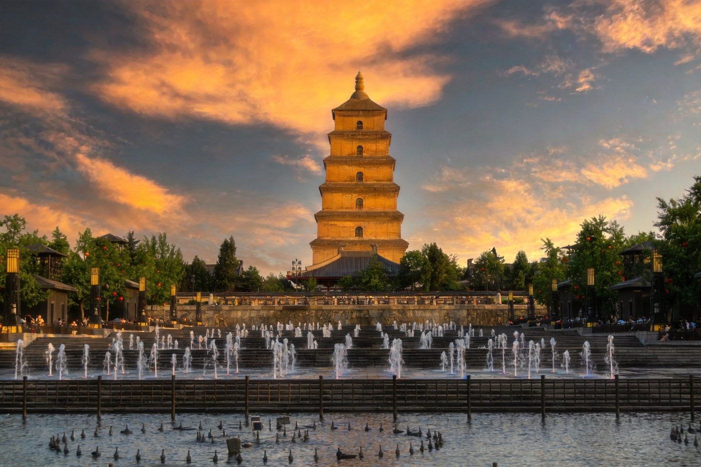 西安大雁塔：唐代方形砖塔耸立傍晚天空下，塔身暖黄古砖，前有喷泉广场，晚霞渐染天际