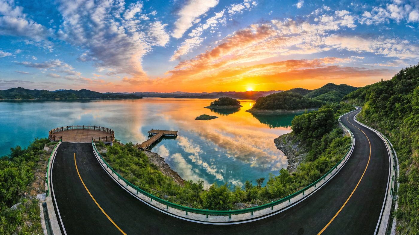 丹江口环库公路碧水连天观景台，柏油路蜿蜒在碧湖与青山之间，木质观景平台延伸入水，夕阳染红天际